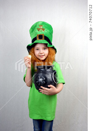 Adorable seven years old girl dressed in green, in leprechaun's hat, holds cast iron pot, full of golden treasure, showing off golden coin with four-petal clover leaf. Saint Patricks Day 74070712