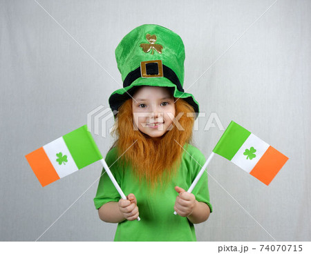 Saint Patricks Dayc elebration. Pretty smiling small girl with decorative red beard in green T-shirt and leprechaun's hat, with flags of Ireland in her hands 74070715
