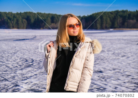 Young female smiling and looking away in sunglasses a snow landscape. Happy hipster girl with hip hop warm winter clothes. Staying on frozen river, lake. 74070802