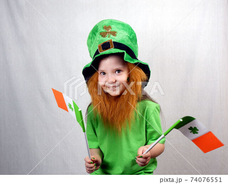Saint Patricks Day holiday. Portrait of adorable smiling girl with decorative red beard, green clover leaf on her cheek and leprechaun hat, holds flags of Ireland in her hands 74076551