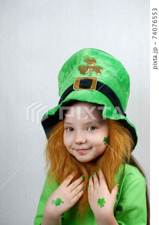 Saint Patricks Day. Endearing smiling small girl with decorative red beard, green shamrok leaf on her cheek and leprechaun hat, raised her hands with green nails and clover leaves to beard 74076553