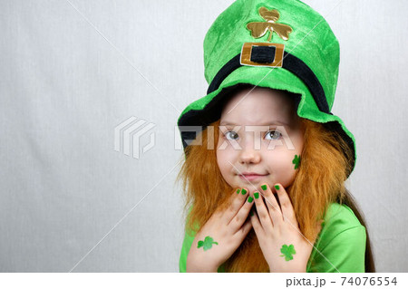 Saint Patricks Day celebration. Nice pensive little girl with decorative red beard, green shamrok leaf on her cheek and leprechaun hat, raised her hands with green nails and clover leaves to her face Saint Patricks Day celebration. Nice pensive little girl with decorative red beard, green shamrok leaf on her cheek and leprechaun hat, raised her hands with green nails and clover leaves to her face 74076554