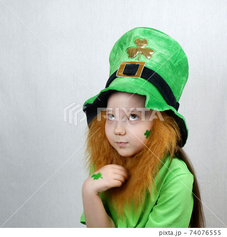 Saint Patricks Day holiday. Portrait of adorable pensive girl with clover leaf on her cheek and hand, decorative red beard in green clothes and leprechaun hat 74076555