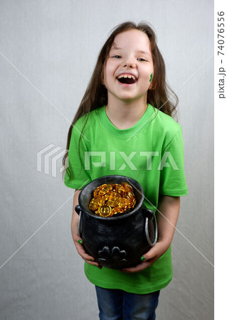 Laughting cute seven years old girl with green shamrok leaf on her cheek showing off  cast iron pot, full of leprechaun gold coins. Saint Patricks Day celebration 74076556