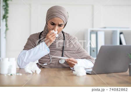 Sick muslim woman doctor sneezing nose and looking at thermometer Sick muslim woman doctor sneezing nose and looking at thermometer 74082862