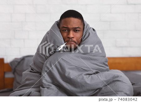 Ill African Guy Holding Thermometer In Mouth Sitting In Bedroom 74083042
