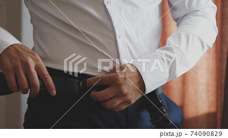 A smart man puts a belt on his pants with a white shirt to keep the pants on his hips. Getting ready early in the morning for work in a room at home. Close-up 74090829