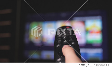 Woman hand selects internet tv channels with remote control, close-up. Person controls TV using a modern remote control. Girl watches smart TV and uses black remote control. Blurry tv scrolls pages 74090831