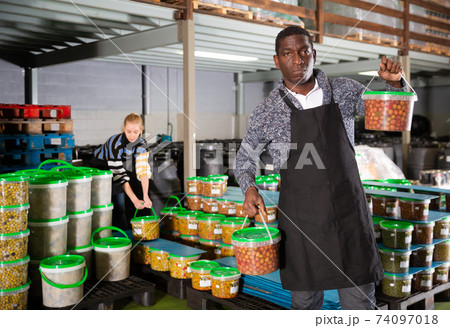 Afro-american male worker stocks plastic containers and cans with olives in warehouse Afro-american male worker stocks plastic containers and cans with olives in warehouse 74097018