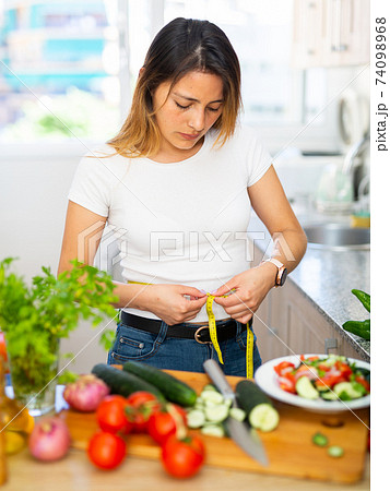 Upset housewife measures waist before eating vegetable salad Upset housewife measures waist before eating vegetable salad 74098968