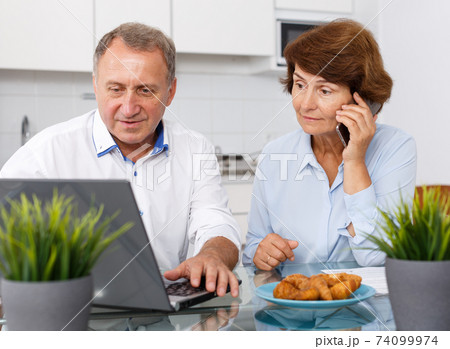 Smiling mature family couple with phone using laptop at kitchen table Smiling mature family couple with phone using laptop at kitchen table 74099974
