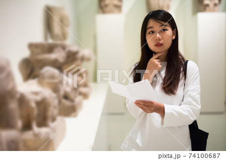 Chinese woman visitor with guide book looking at exhibition in museum Chinese woman visitor with guide book looking at exhibition in museum 74100687