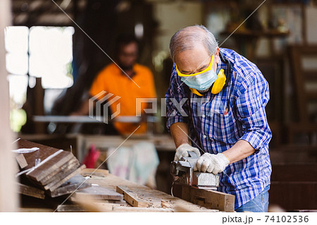 asian senior man carpenter wear protective mask dust and headphone working with electric planer. asian senior man carpenter wear protective mask dust and headphone working with electric planer. 74102536