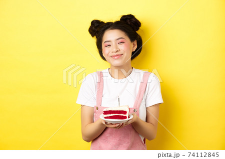 Happy asian birthday girl with bright makeup, blowing candle on cake, making wish, standing on yellow background 74112845