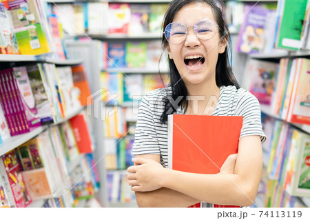 Smiling asian child girl wearing glasses holding a book on her arms,female student standing feel enjoy in book store,happy woman is laughing, relax by reading books,concept of development,education Smiling asian child girl wearing glasses holding a book on her arms,female student standing feel enjoy in book store,happy woman is laughing, relax by reading books,concept of development,education 74113119