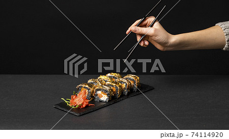 Hand with black chopsticks wants to take custom sushi roll with cuttlefish ink, salmon, cream cheese, pepper pumpkin, eel and edible gold leaf with ginger, wasabi on a square plate on black table. 74114920
