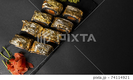 Custom sushi roll with nori, cuttlefish ink, fresh salmon, cream cheese, pepper pumpkin, eel, unagi sauce and edible gold leaf with red ginger, wasabi on a square plate on black table and background. Custom sushi roll with nori, cuttlefish ink, fresh salmon, cream cheese, pepper pumpkin, eel, unagi sauce and edible gold leaf with red ginger, wasabi on a square plate on black table and background. 74114921