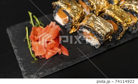Custom sushi roll with nori, cuttlefish ink, fresh salmon, cream cheese, pepper pumpkin, eel, unagi sauce and edible gold leaf with red ginger, wasabi on a square plate on black table and background. Custom sushi roll with nori, cuttlefish ink, fresh salmon, cream cheese, pepper pumpkin, eel, unagi sauce and edible gold leaf with red ginger, wasabi on a square plate on black table and background. 74114922