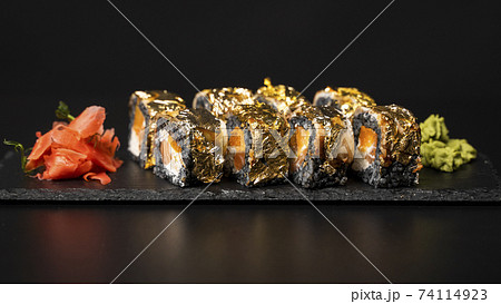 Custom sushi roll with nori, cuttlefish ink, fresh salmon, cream cheese, pepper pumpkin, eel, unagi sauce and edible gold leaf with red ginger, wasabi on a square plate on black table and background. Custom sushi roll with nori, cuttlefish ink, fresh salmon, cream cheese, pepper pumpkin, eel, unagi sauce and edible gold leaf with red ginger, wasabi on a square plate on black table and background. 74114923