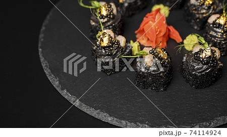 Sushi roll with nori, black rice, crab meat, cucumber, avocado, smoked salmon mousse, oar caviar, masago, shrimp cocktail and edible gold leaf with red ginger and wasabi on black table and background. Sushi roll with nori, black rice, crab meat, cucumber, avocado, smoked salmon mousse, oar caviar, masago, shrimp cocktail and edible gold leaf with red ginger and wasabi on black table and background. 74114924