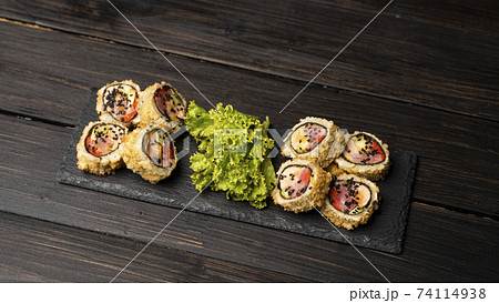 Custom sushi roll in tempura with nori, fresh salmon, tuna, avocado, masago caviar, drizzled with pineapple sauce with salad pouring as decoration on a black plate on a wooden table and background. 74114938