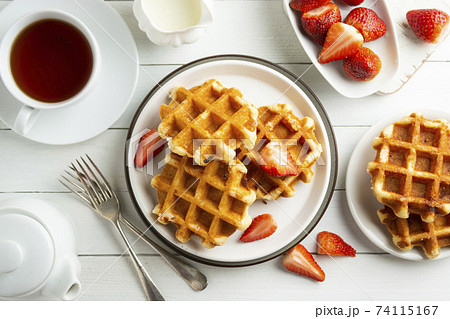 Delicious breakfast. Belgian waffles with honey and strawberries. Cup of tea white wooden background, top view. Delicious breakfast. Belgian waffles with honey and strawberries. Cup of tea white wooden background, top view. 74115167