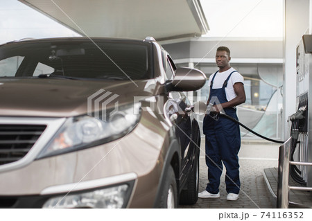 Young handsome African male worker at gas station refueling client's car with petrol 74116352