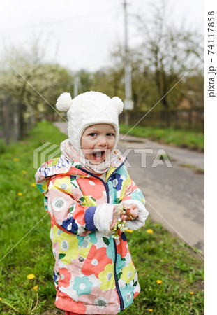 a little girl in a bright jacket is laughing happily outside in the spring and holding a branch with spring white flowers. Children's happiness. a little girl in a bright jacket is laughing happily outside in the spring and holding a branch with spring white flowers. Children's happiness. 74117858