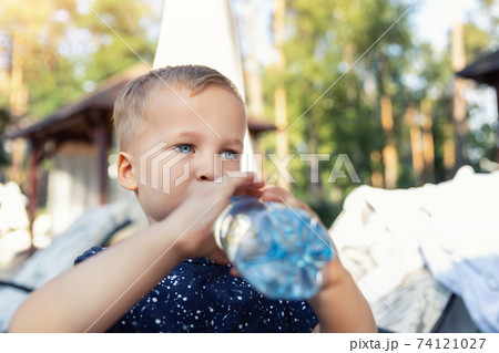 Portrait of cute playful little caucasian blond thirsty toddler boy kid holding plastic bottle and drinking water on hot summer day outdoors. Children heat dehydration danger awareness 74121027