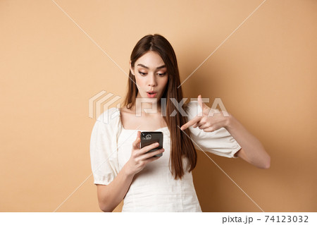 Exciting news on phone. Surprised cute woman reading online promo on screen, pointing at smartphone and say wow amazed, standing on beige background Exciting news on phone. Surprised cute woman reading online promo on screen, pointing at smartphone and say wow amazed, standing on beige background 74123032
