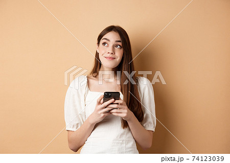Romantic girl thinking with phone, looking aside and smiling dreamy, using dating app on smartphone, standing on beige background Romantic girl thinking with phone, looking aside and smiling dreamy, using dating app on smartphone, standing on beige background 74123039