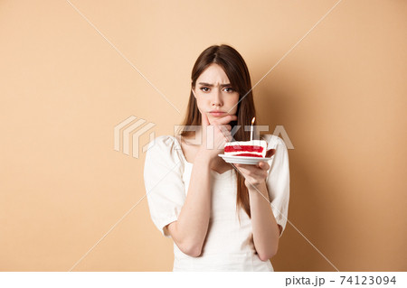 Thoughtful birthday girl frowning and thinking of wish, holding cake with candle, standing on beige background Thoughtful birthday girl frowning and thinking of wish, holding cake with candle, standing on beige background 74123094