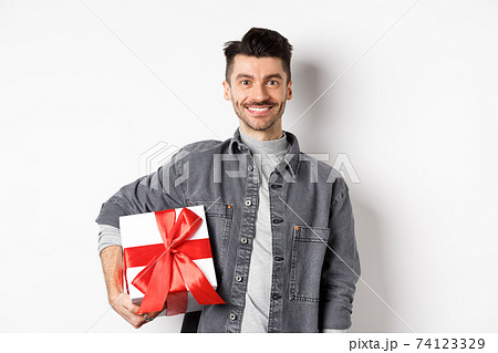 Happy Valentines day. Smiling handsome man bring gift on romantic date, holding box with present and looking romantic at camera, white background Happy Valentines day. Smiling handsome man bring gift on romantic date, holding box with present and looking romantic at camera, white background 74123329