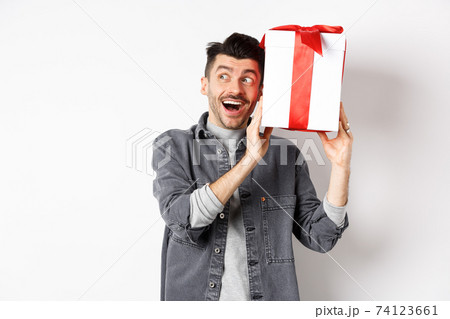Happy boyfriend shaking his gift to guess what inside, receive surprise present on Valentines day, smiling and looking aside cheerful, standing on white background Happy boyfriend shaking his gift to guess what inside, receive surprise present on Valentines day, smiling and looking aside cheerful, standing on white background 74123661