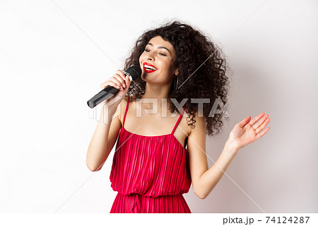 Elegant curly-haired woman in red dress singing in microphone, looking aside and smiling happy, standing on white background 74124287