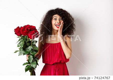 Excited curly woman in red dress, receive bouquet of roses and look surprise, rejoicing from romantic gift, standing on white background Excited curly woman in red dress, receive bouquet of roses and look surprise, rejoicing from romantic gift, standing on white background 74124767