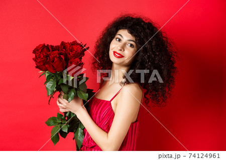 Happy beautiful woman in dress, holding flowers and smiling romantic, standing against red background 74124961