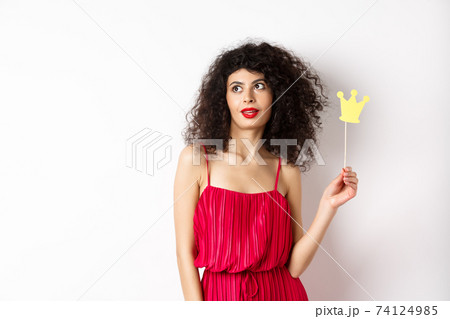 Stylish woman with curly hair in red dress, holding queen crown on stick and looking aside, standing on white background 74124985