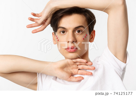 People, lgbtq and beauty concept. Close-up of beautiful androgynous male model with glitter on face, posing sensual at camera, standing over white background People, lgbtq and beauty concept. Close-up of beautiful androgynous male model with glitter on face, posing sensual at camera, standing over white background 74125177