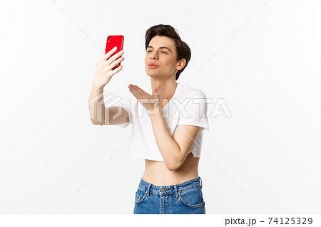 Beautiful gay man in crop top sending air kiss at phone camera, taking selfie or video chat on smartphone, standing over white background 74125329