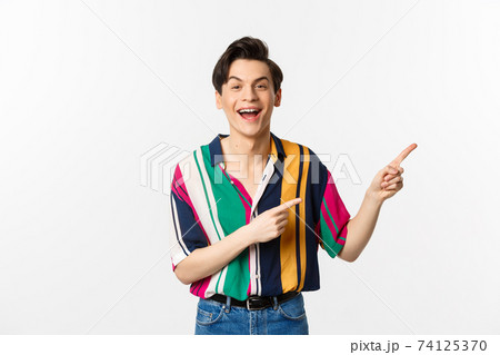 Happy gay man laughing and smiling, pointing fingers right at promo offer, standing over white background 74125370