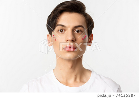 People, lgbtq and beauty concept. Close-up of beautiful androgynous man with glitter on face, looking at camera, standing over white background 74125587