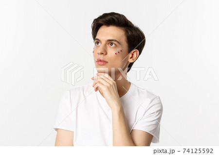 People, lgbtq community and lifestyle concept. Beautiful androgynous man with glitter on face, looking at upper left corner sensual, standing over white background 74125592