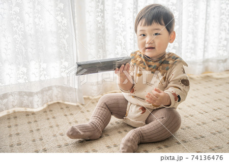 窓辺でお座りをしてリモコンで遊んでいる赤ちゃん 74136476