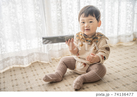 窓辺でお座りをしてリモコンで遊んでいる赤ちゃん 74136477