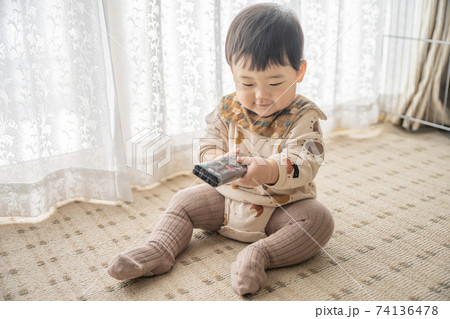 窓辺でお座りをしてリモコンで遊んでいる赤ちゃん 74136478