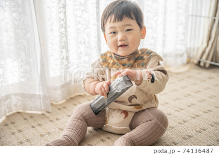 窓辺でお座りをしてリモコンで遊んでいる赤ちゃん 74136487
