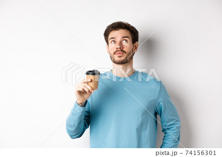 Image of young man drinking takeaway coffee and looking up with thoughtful face, tasting new flavour, standing over white background 74156301