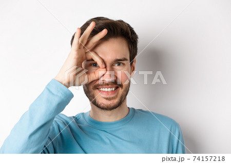 Close up of happy man looking through okay sign and smiling, approve and like something good, standing over white background Close up of happy man looking through okay sign and smiling, approve and like something good, standing over white background 74157218
