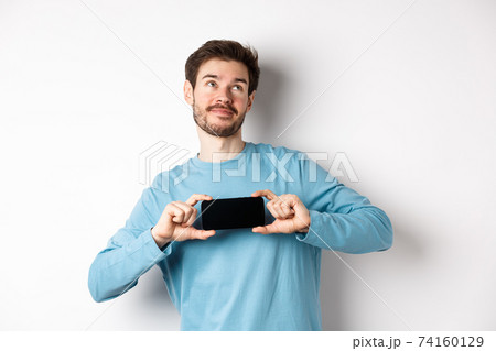 E-commerce and shopping concept. Smiling young man showing mobile screen and dreaming about something, looking at upper right corner nostalgic, standing over white background 74160129